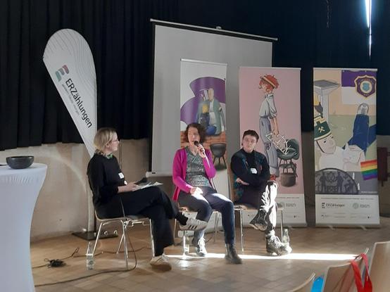 Podium mit drei Frauen, im Hintergrund drei Roll-Ups mit Zeichnungen, die erzgebirgische Motive und Traditionen neu interpretieren.
