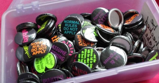 A boxful of one-inch black button badges with colourful text.  They don't yet have their pins installed on the backs.  Among the visible designs are
"Normal rules do not apply"
"Assume nothing"
"They"
"Visibility", with the "Bi" in the middle in a wider font so that it's sort of like "bi visibility".
"... and the princess fell in love with the other two princesses, and they all lived happily ever after"