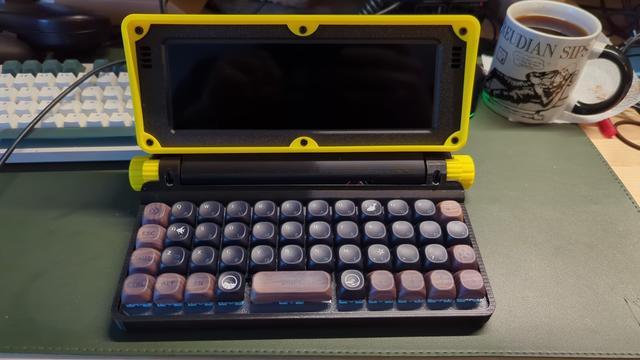A MicroJournal Rev2 writing-deck device in yellow/black plastic with black and wood grain keys in matrix arrangement (no staggering). It sits on a large green mat in front of a normal mechanical keyboard and a coffee cup that says "Freudian Sips"