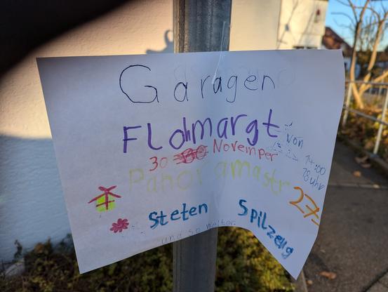 Ein handgeschriebener Zettel in bunter Kinderschrift, der auf den Garagenflohmarkt aufmerksam macht, klebt an einem Schildermast.