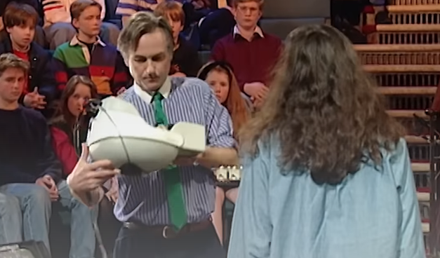 Frame de um vídeo de aula pública com Richard Dawkins, homem com camisa listrada azul e branca (estilo Bananas de Pijamas) e uma gravata verde, está segurando um capacete de realidade virtual. Ao fundo, jovens na plateia. No primeiro plano, uma garota de cabelos longos e jaqueta jeans de costas.