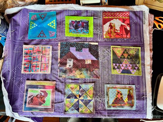 Fabric is arranged on a purple base with intricate circuit-like patterns, and various colorful square patches are laid onto it. Each square features unique designs, such as geometric patterns, abstract art, and imagery of animals or houses. Tools like scissors and other crafting supplies are visible in the background.