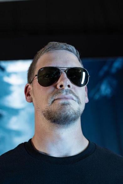 Portraitfoto von Frederick. Er trägt ein schwarzes Shirt und Sonnenbrille vor einem blauen Winterwald