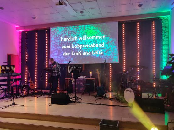 Chorraum einer modernen Kirche, stimmungsvoll beleuchtet, mit aufgebautem Band-Equipment. Auf der Leinwand ein grün-blaues Muster, darauf der Text "Herzlich willkommen zum Lobpreisabend der EmK und LKG"