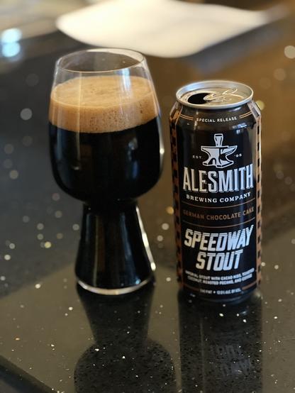 A stout glass full of dark beer with a thick tan head. The pint can is black with white print, the AleSmith anvil logo is up top, the finish line checkerboard is just barely visible in brown to the right