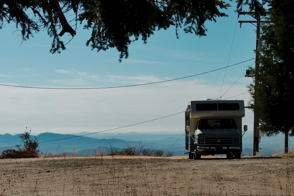 A photo of a camper van sitting on flat, level, bare dirt. Behind it, wires frame a band of cloud above a mountainous expanse that is mostly below the point of view. Above and to its right, evergreens and a telephone pole frame the image.