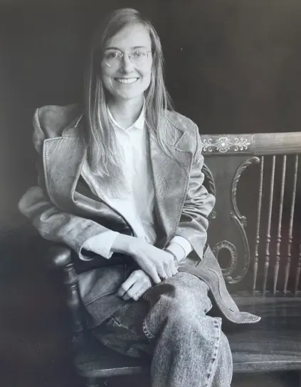 A woman with long, light brown hair, sitting in a ornate wooden chair. The woman is wearing glasses with a wide smile, and is wearing a dress shirt, jacket, and blue jeans. Her hands are folded on her lap.