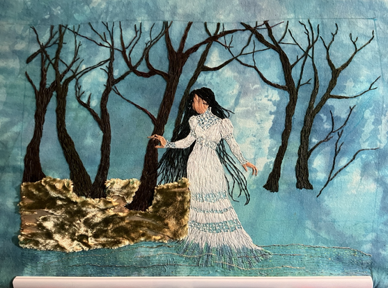 An embroidered panel in progress of La Llorona in a white gown standing in the edge of a river, with a background of mesquite trees growing from a tan and olive mossy velvet ground.