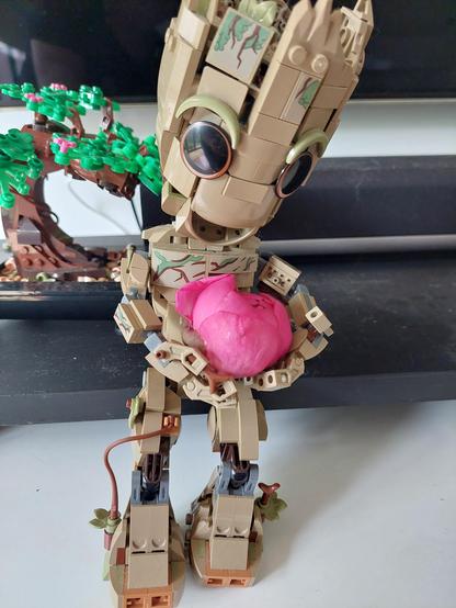 A Lego Groot is holding a large deep pink flower bud in his hands. His head is tilted to the side slightly and he is looking down at the flower bud.