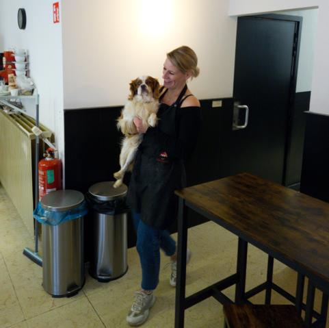 A woman is walking from the back into her store with her dog, smiling big time.