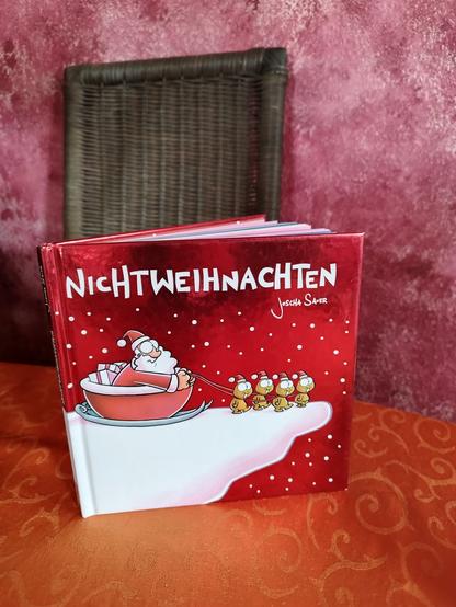 ein rotes kleines Buch mit glänzendem Einband steht leicht geöffnet auf einem Tisch .. der Titel:
'NICHTWEIHNACHTEN' von Joscha Sauer