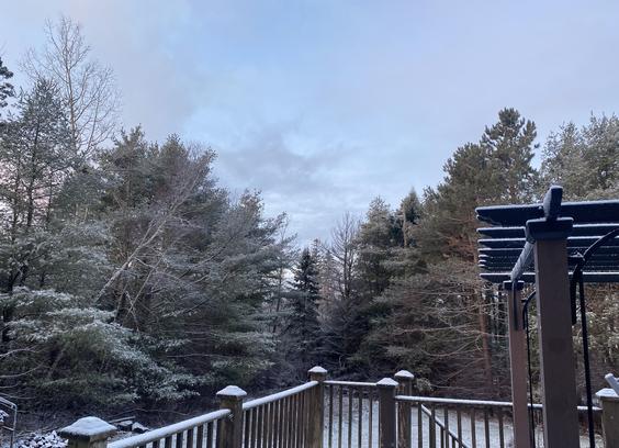 Deck rails and a steel pergola with small dusting of snow. Grass and trees behind can also be seen with small amount of snow. Pretty blue sky with wispy clouds above.