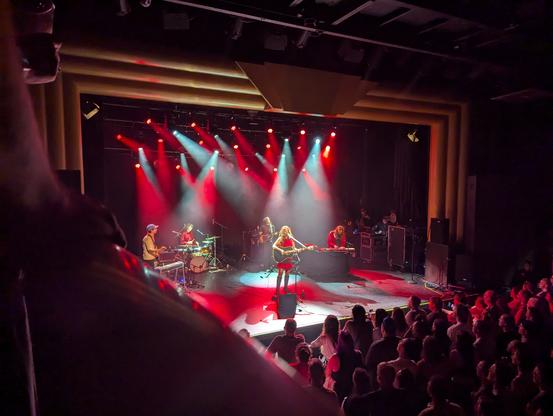 Photograph of a concert stage with a band playing under red and turquoise lights. The photo is from a balcony along the left hand side of the hall. Katie Crutchfield is centre stage with a black acoustic guitar. The four other musicians in the band are arranged behind her.