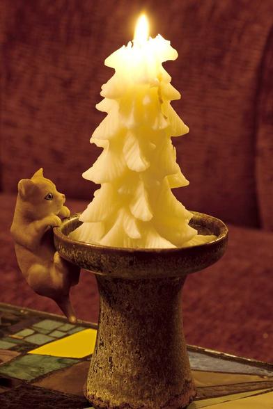 Eine brennende, tannenbaum-förmige Kerze auf einem Keramik-Kerzenständer, an dem auf der linken Seite eine kleine Katze aus Wachs hängt.