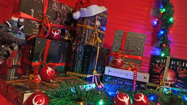 A festive arrangement of boxes for all of the retail releases of the Quake series, ranging from the simple box art of Quake with its iconic rusted 'Q' logo, to Quake 3 Arena's shiny metallic tin complete with greebles and a fancy re-imagining of the Quake logo in embossed red. Garlands, ornaments, and Christmas lights are scattered throughout the scene, the ornaments having little Quake symbols painted on them, and each box is wrapped with shiny red or golden bows; one box even has a Santa hat draped atop of it. A small figurine of a character from Quake Champions stands with arms folded and hands resting on a rocket launcher, and he, too, has a Santa hat on his head.