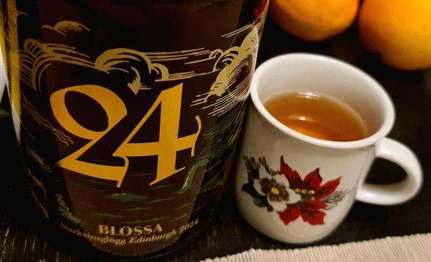 A bottle to the left with "24" in big numbers, under is the brand "Blossa". To the right is a small cup with two flowers on, and is containing orange liquid (mulled white wine). In the background are parts of two oranges