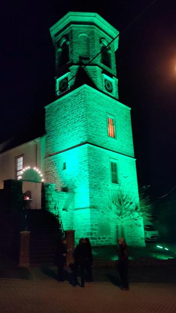 Die Dorfkirche Bucha ist in grünes Licht getaucht. Über dem Eingang zum Kirchgarten ist ein beleuchteter Schwibbogen zu sehen.