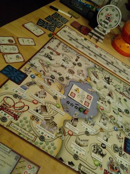 Schräge Aufnahme eines Holztischs mit großem Spielplan in Form eines Freizeitparkplans. Die Attraktionen, Shows, Restaurants, Shops und Info-Punkte besitzen jeweils ein Feld, welche über verschlungene Wege miteinander verbunden sind. Links vom Plan liegen verschiedene Karten, teils verdeckt. Hinter dem Plan steht ein ca. 20 cm hohes Glücksrad, zusammengesteckt aus Pappe. In der Mitte befindet sich ein See mit einer aufgedeckten Karte, darunter Sterne mit aufsteigenden Punkten, auf den beiden rechten mit den höchsten Punktzahlen stehen zwei Meeple.