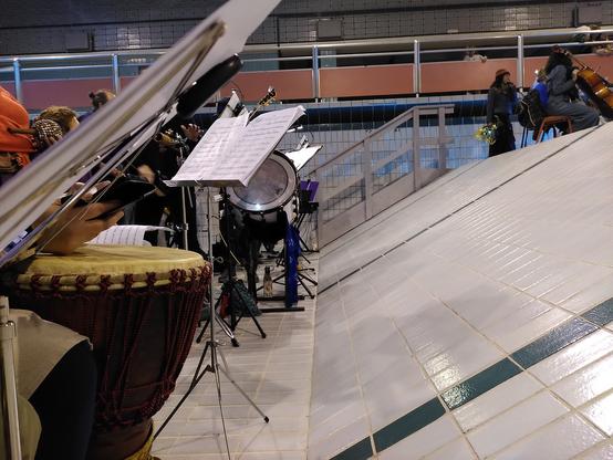 Vue prise du fond d'une piscine sans eau, au pied du plan incliné. On voit des pupitres des instruments et des musicien•ne•s