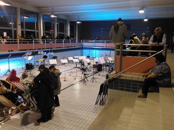 Pupitres et instruments au fond d'une piscine sans eau. Public debout sur le bord et installé dans des transats côté petit bain. Éclairage de scène bleu. La photo est prise du bord côté petit bain.