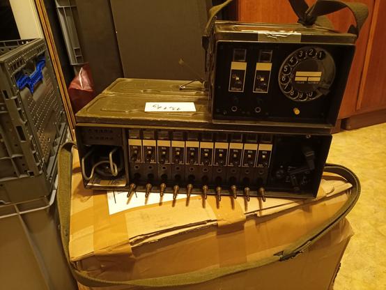 Auf einem Karton steht eine Feldtelefon Vermittlung mit klinken Steckern und hebeln dort drüber steht der dazugehörige amts Zusatz mit Wählscheibe.