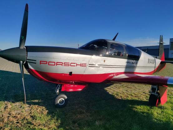 A sleek black, grey and red single-engine light aircraft with a black nose and three-bladed propeller, photographed at close distance from the front left, in bright sunlight. It has a prominent red "PORSCHE" decal on the engine cowling. It sits on grass; in the background are a paved surface and some hangars. The sky is bright blue with a few thin clouds near the horizon.