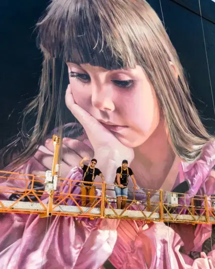 The image shows a large mural of a young girl with long brown hair, wearing a pink garment. Her expression is contemplative, with her chin resting on her hand. 

In front of the mural, the two painters are standing on a suspended platform. They are wearing safety harnesses and casual clothing. One of the men is waving at the camera. 

The mural is highly detailed and realistic, covering a large section of a building wall.