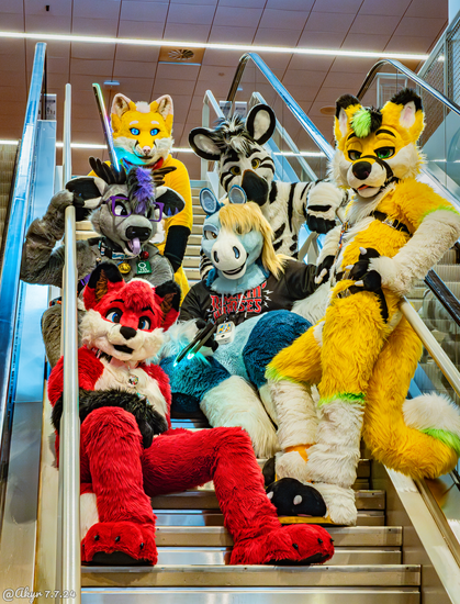 This is a group photo from Anthrocon 2024 of six fursuiters sitting on a stairway between two escalators. In the center is a blue and white pegasus wearing a black baseball jersey, and going around him clockwise are a gray and purple deer, a yellow and pink fox with a glowing LED sword, a white and black zebra, a yellow and green fox-husky, and a red and white fox.