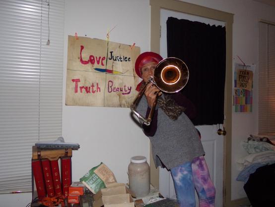 Love Justice Truth Beauty on a veneer poster sits on a wall behind a woman wearing a red knit hat calico infinity scarf grey puffy turtle neck sweater and purple tie dye leggings blowing a copper-belled bass trombone 4 of 5 volumes of 2023 Bigelow essay winners gifted to me this year from New Thinking Allowed