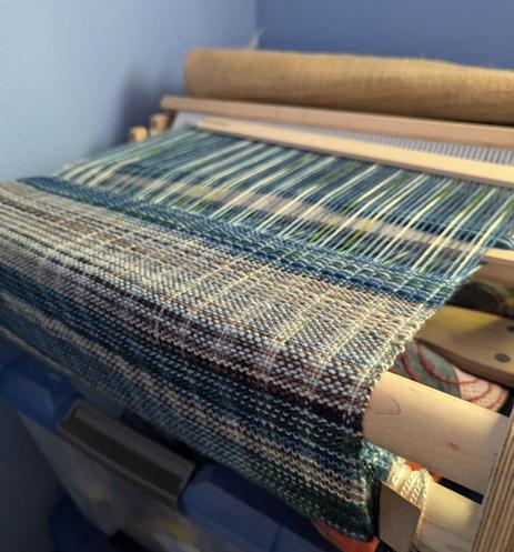 A weaving in progress on a rigid heddle loom.  it uses two colours of variegated yarn, one grey/brown and the other blue/green.  It's a sampler showing multiple types of pattern that can be made with a pick up stick.