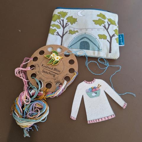 A small embroidery kit including a nice round yarn holder, a piece of felt pre-printed with an embroidery pattern (a sweater with a sheep and yarn on it) and a small bag with a camping motf.