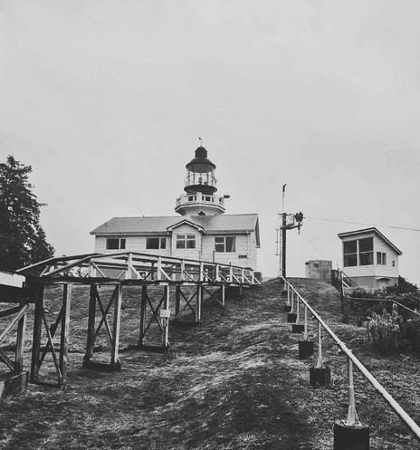 A historic lighthouse stands atop a hill, featuring a white building with a conical lantern. A very old & rotting wooden walkway leads to the lighthouse, flanked by a grassy area & trees. Two smaller structures are visible nearby. The sky is overcast. Black and white photo.