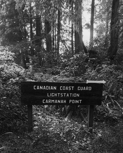 A wooden sign with white arrow pointing to the right. It displays the words "Canadian Coast Guard Lightstation Carmanah Point," indicating the location. The sign is set against a backdrop of dense trees & foliage in the forest. Black and white photo.