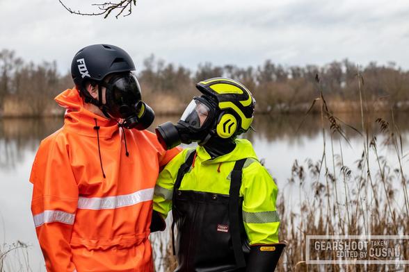 On a gloomy day, in front of a lake that is surrounded by reeds, the fox and the cat-dog stand arm in arm, looking at each other's eyes. The fox wears an neon-orange raingear one-piece with reflective stripes, a black gas mask and a black FOX helmet. Adensy wears neon-yellow raingear, half covered by black chest waders, a gas mask and a neon-yellow helmet with ear protectors.