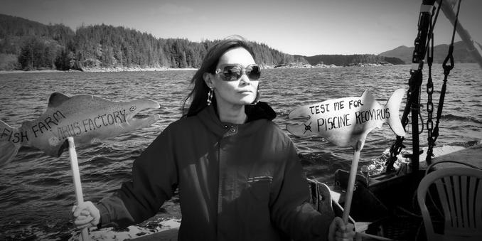 Me, standing on a large fishing boat, looking solemn while holding two signs shaped like salmon. One sign reads "FISH FARM VIRUS FACTORY," and the other says "TEST ME FOR PSCINE REOVIRUS." The scene is in black and white, with water and a forested shoreline visible in the background, creating an atmosphere of protest against fish farming practices.