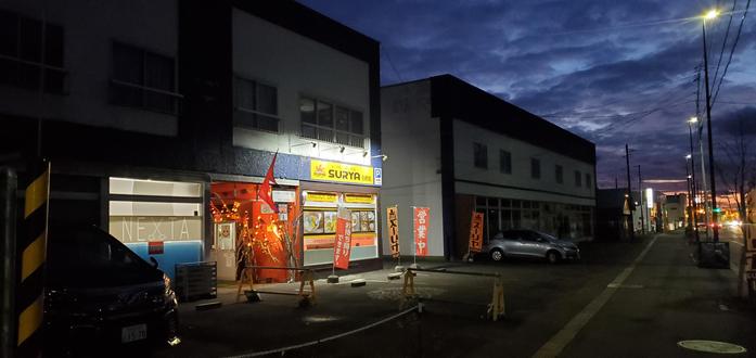 Exterior of the restaurant Surya on the main street of a small town in Hokkaido. It is just after sunset and the cloud covering suggests later rain. There are festive lights and Nepali decor in the alleyway beside the restaurant. The other buildings are shrouded in late afternoon darkness.