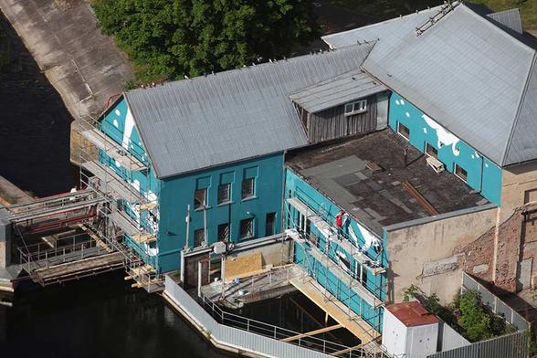 An aerial view of the turquoise mural by artist Ray Bartkus in Marijampolė, Lithuania, painted on the side of a building adjacent to a canal. The image shows the mural in progress, with scaffolding and workers visible as they paint the upside-down design. The mural is designed to reveal its full meaning when reflected in the water, creating an upright scene of swimmers, rowers, and swans.