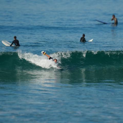 Bodysurfer with one arm extended with a handplane riding the wave, with other arm extended behind him as a wave breaks. Surfers with surfboards are waiting for waves further out in the surf.