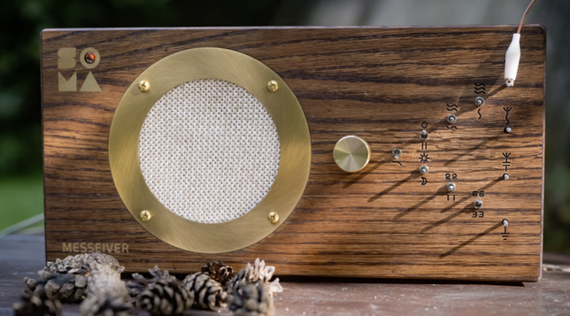 The SOMA MESSSEIVER: a beautiful wooden electronic device with a retro-futuristic aesthetic. There's a circular brass speaker grille on the left side, and a small round brass knob and various little switches and buttons labelled by symbolic engravings that look like alchemical glyphs and ogham runes. The words SOMA and MESSEIVER are engraved on the wooden casing. Some pinecones are scattered in the foreground.