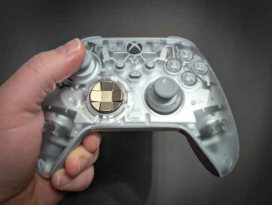 A hand is holding a translucent Xbox controller. The controller has a frosted, semi-transparent casing that reveals some of the internal components. The buttons and joysticks are light gray, and the directional pad is metallic gold. The Xbox logo is visible at the top center of the controller.