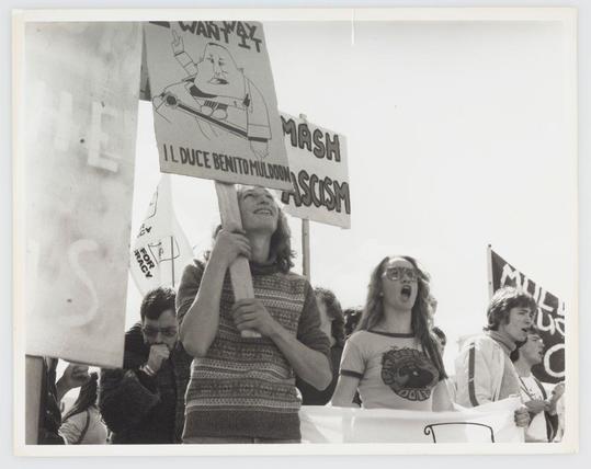 A black and white photograph depicting a group of protesters, some holding signs with messages. The most prominent sign reads "I Duce Benito Muldoon," which is associated with the political figure Pinochet, suggesting opposition to his rule in Chile during that time period. Other visible text on various placards includes phrases like "MASH" and what seems to be a call for economic reform or government change ("WE WANT A PAY increase"). The protesters appear animated, some singing and others shouting slogans; their expressions convey determination and perhaps frustration.