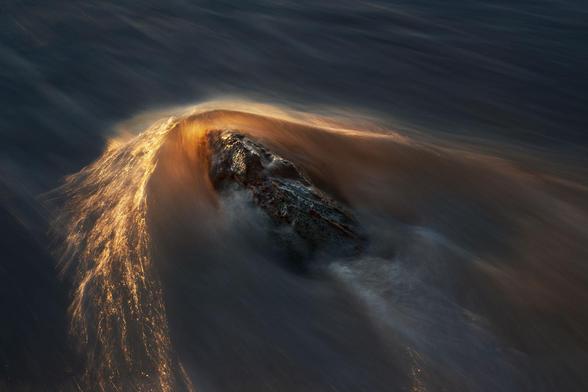 a rock with the water rushing against it. The water is lit by the low sun making it orange. It can also be read as if the rock is rushing through the water.
