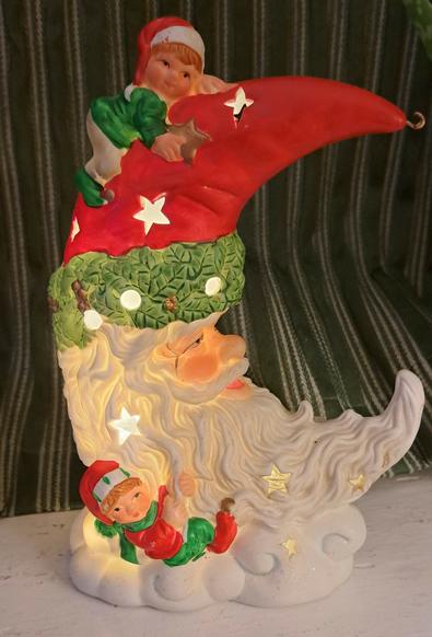 A ceramic crescent moon which looks like Santa. There are two elves. One is on his hat, the other clinging onto his beard. The moon is pierced with stars and the light inside can be seen through the stars.