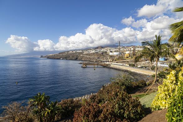 A scene of almost tropical brightness and colouring. We're on a hill, part of a wide promontory jutting out from the land which we can see below and to the right in lower hills curving around a wide bay of deep blue water. From this distance the water appears nearly flat, barely rippled with blue shadows and reflecting the banks of puffy white clouds on the horizon. This hill we're on is part of a tropical garden specialising in palms, a few of which are framing the view on the right near us, but in front it's mostly smaller plants and bushes in shades of bright yellow-greens and darker red-greens with well-trimmed grass visible between. The land to the right contrasts starkly, filled as it is with a sprawling oil refinery, but one with bright colours on silos and towers lifting the industrialised sight to something seemingly more friendly and suitable for the scene. It feels very warm with just a hint of refreshing sea air coming up from below.