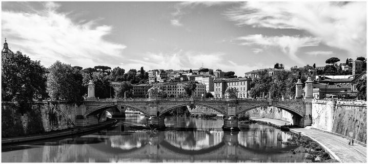 Black and white image of a historic bridge spanning a river, surrounded by trees and buildings in a scenic urban landscape. The sky is partly cloudy, reflecting on the water.