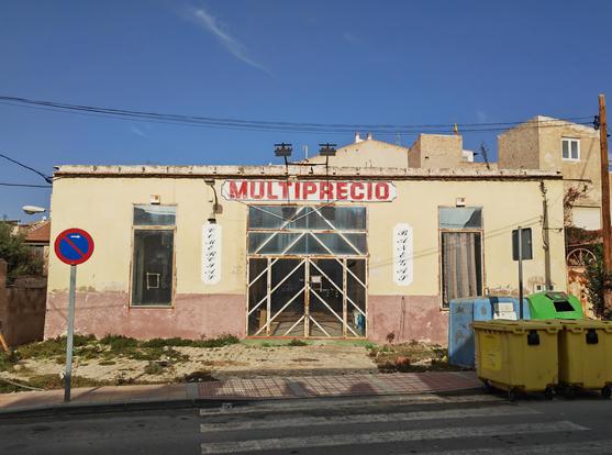 An abandoned shop. It is a plain rectangular box with a big rectangular opening in the middle and a tall rectangular opening on each side, evenly distant from the door.

The door and surround are glass, held together by a welded frame of iron sections, vertical and diagonal, forming a child's storybook sentry post. 

The sign over the door says 'Multiprecio', in capital letters