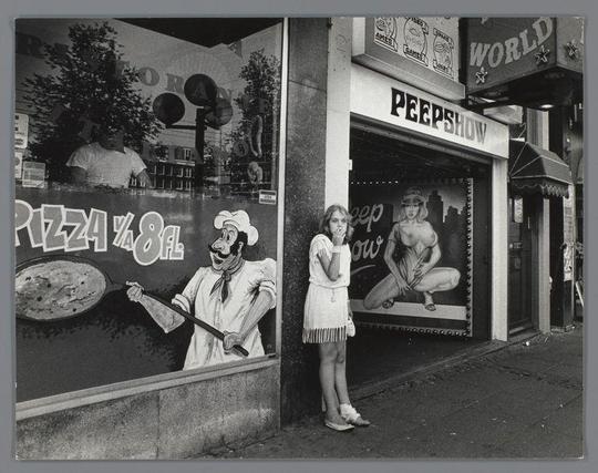 The image depicts a monochrome street scene featuring an advertisement for "Peepshow" with large, bold text. The ad includes images of food items and suggestive imagery, such as a person holding their chest in a provocative manner.

In front of the store entrance stands a young woman wearing a white dress, observing something to her side while resting her chin on one hand. She appears contemplative or possibly startled by an unseen presence nearby. Her attire is casual, with sandals on her feet and what looks like bandages wrapped around both ankles. The street setting has other storefronts in the background, contributing to an urban atmosphere.

The photo captures a candid moment that evokes curiosity about the context of this encounter between the woman and whatever she's reacting to off-camera.