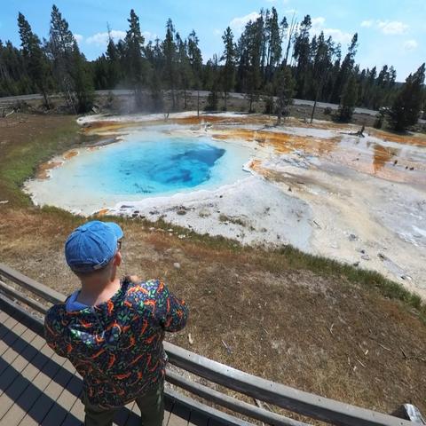 Person standing on a boardwalk, observing a nearby hot spring in Yellowstone National Park.