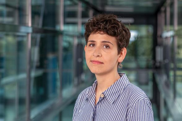 Photo of a person with shorter, curly brown hair wearing a blue and white striped shirt. In the background, glass fronts with dark gray struts can be seen out of focus on the left and right. Picture: sevens[+]maltry