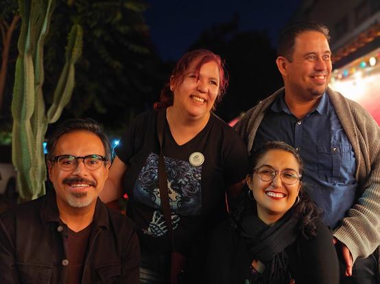 Dos hombres y dos mujeres posan, dos sentados y dos de pie, tras una mesa al aire libre, entre un cacto en maceta y un toldo iluminado.